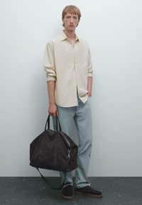 Lichtbeige knoopshirt, lichtblauwe jeans en een donkerbruine suède duffeltas met handvatten, gedragen met donkere schoenen tegen een neutrale achtergrond.