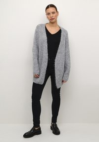 Lichtgrijze gebreide cardigan met een open voorkant, lange mouwen en een pluizige textuur, gecombineerd met zwarte strakke broeken en zwarte instappers.