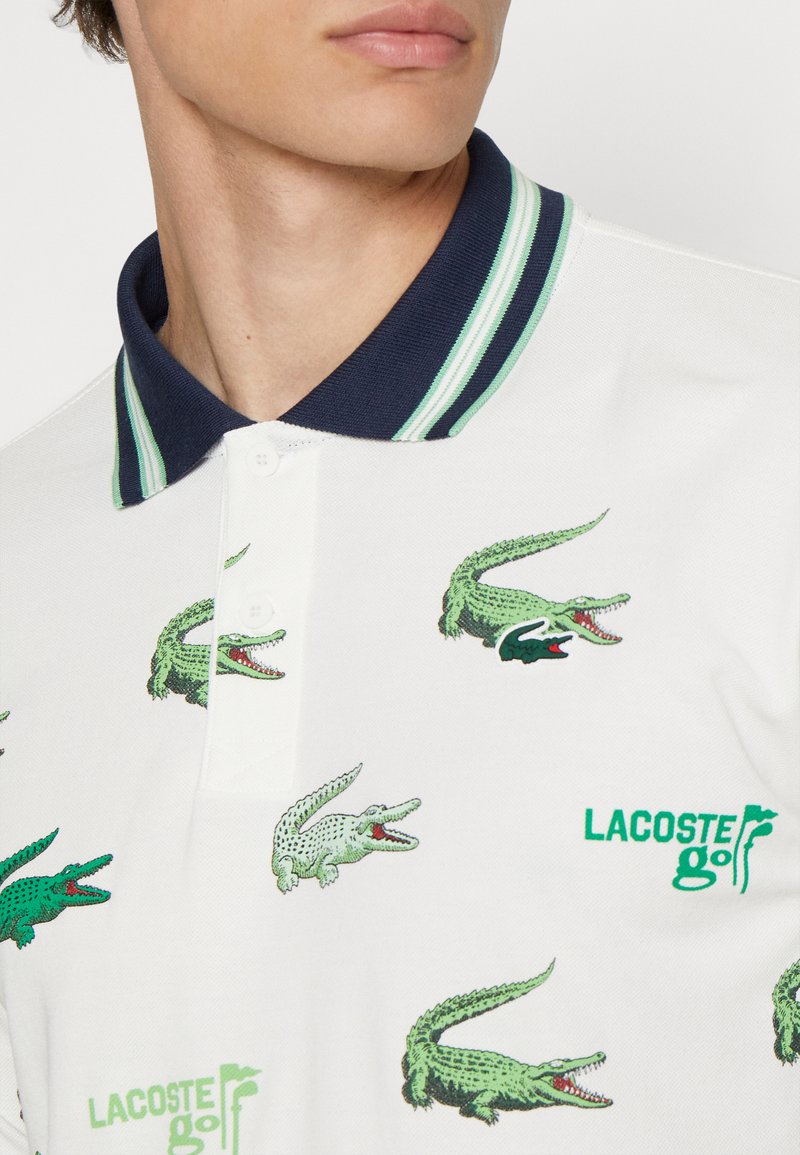 Polo blanc avec un col rayé bleu marine, comportant des graphiques de crocodile verts et le texte "LACOSTE golf" en vert.