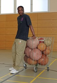 Black graphic T-shirt, khaki pants with buttons on sides, white sneakers, leaning on a metal cart filled with various basketballs, indoor setting.