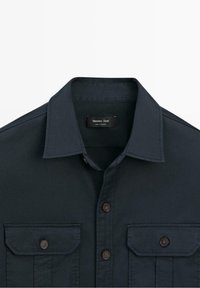 Chemise bleu foncé à boutons, avec deux poches poitrine, rabats boutonnés et col pointu, avec étiquette de marque à l'intérieur du col.