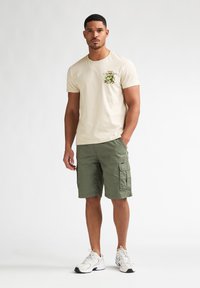 Beiges Baumwoll-T-Shirt mit floralem Grafik-Taschendetail, kombiniert mit olivgrünen Cargo-Shorts und weißen Sportschuhen.