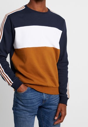 Hombre vistiendo una sudadera de bloques de color con secciones en azul marino, blanco y marrón, y detalles a rayas en las mangas, combinada con vaqueros azules.