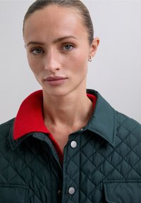 Chaqueta acolchada verde con un cuello acanalado rojo, que presenta botones a presión y un bolsillo frontal. Textura suave y detalle de patrón de diamante.