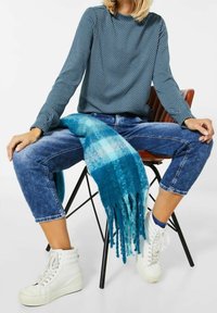 Blå herringbone långärmad topp i kombination med blå jeans. En turkos franssjal hänger över knäna, kompletterad med vita högstövlar.