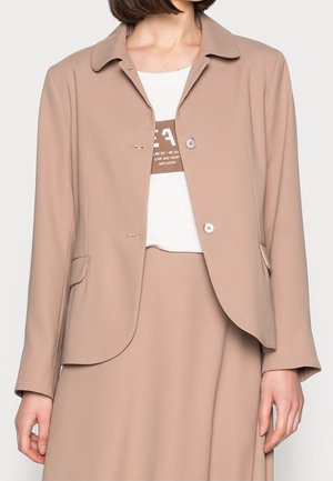 Kvinde iført en beige blazer med knapper over en hvid skjorte med brun tekst, kombineret med en matchende beige nederdel.