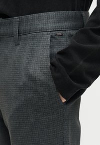 Χέρι στην τσέπη γκρί καρό παντελονιού συνδυασμένου με μαύρο μακρυμάνικο τοπ, που δείχνει την υφή του υφάσματος και μικρή μεταλλική λεπτομέρεια κοντά στην τσέπη.