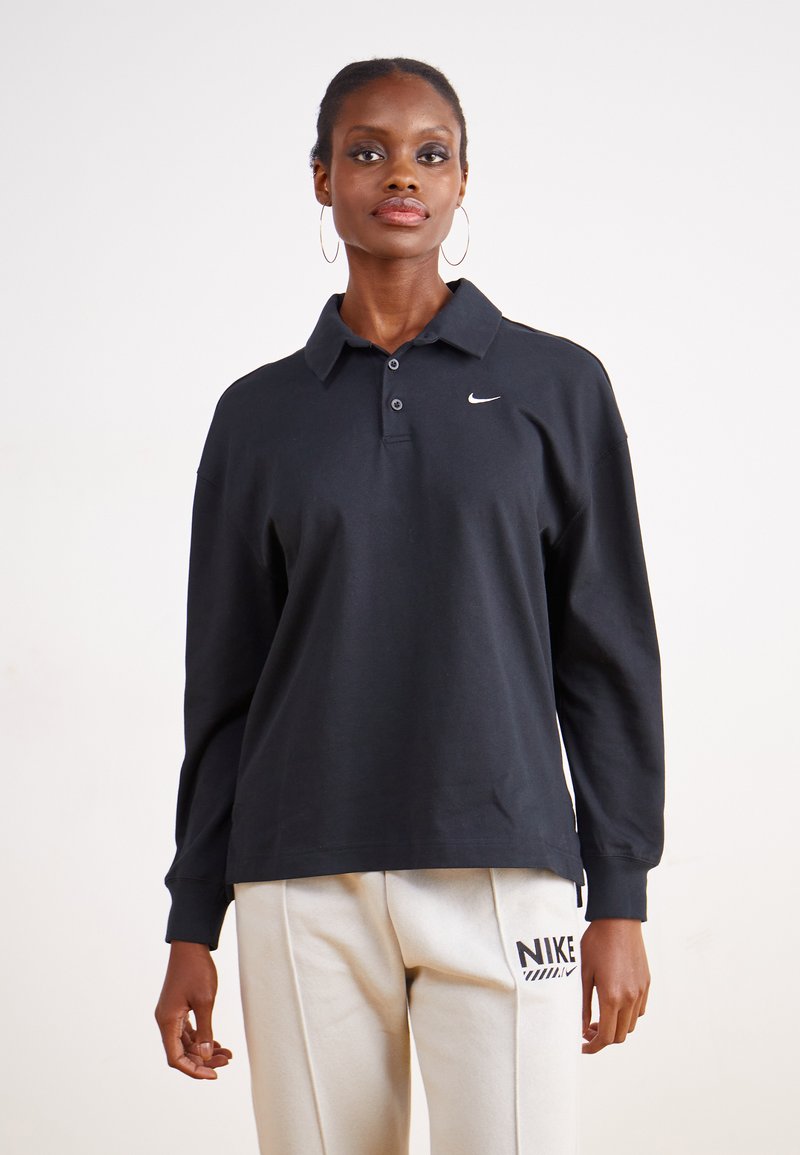 Camisa polo negra de manga larga con cuello abotonado, que presenta un pequeño logo blanco de Nike en el pecho y una textura de tela suave.