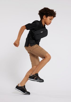 Chemise noire à manches courtes, shorts beiges, baskets noires avec des accents marron, et un sac bandoulière noir, montrés lors d'un mouvement dynamique.