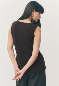 Top senza maniche di colore marrone scuro con una texture a coste, scollatura rotonda e design aderente. Indossato con pantaloni neri, mani unite dietro la schiena.