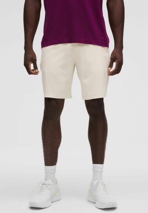 Uomo che indossa pantaloncini color crema, scarpe da ginnastica bianche, calzini bianchi e una maglietta viola scuro, in piedi contro uno sfondo uniforme.