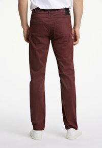 Bordeaux kleurige katoenen broek met een rechte pasvorm, voorzien van twee achterzakken en een logo label op de tailleband. Witte sneakers zichtbaar aan de onderkant.