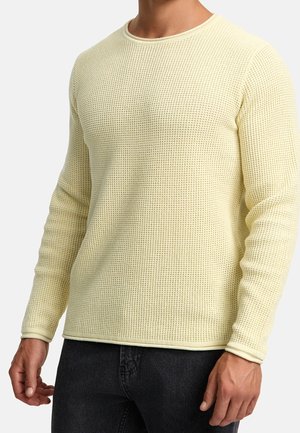 Pull en tricot jaune clair avec un motif texturé. Encolure ronde côtelée et poignets côtelés. Manches longues. Porté avec un jean gris foncé.