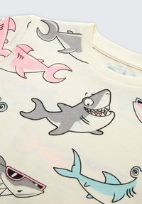 Cremfarbendes T-Shirt mit Cartoon-Hai-Designs in Pink, Grau und Blau. Der Stoff ist weich, hat einen runden Halsausschnitt und kurze Ärmel.