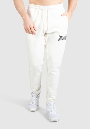 Weiße Jogginghosen mit elastischem Bund, auf der linken Oberschenkelseite mit "SMILDO" in Schwarz bedruckt. Getragen mit weißen Sportschuhen.