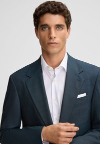Jonge man met krullend bruin haar, gekleed in een donkerblauwe colbert over een wit dress shirt, tegen een eenvoudige lichte achtergrond.