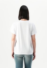 Person med skulderlangt mørkt hår iført en enkel hvit t-skjorte og blå jeans, vendt bort fra en hvit bakgrunn.