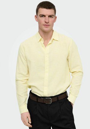 Joven con cabello corto y castaño, vestido con una camisa amarilla clara abotonada y metida dentro de pantalones negros con cinturón marrón, de pie con una mano en el bolsillo.