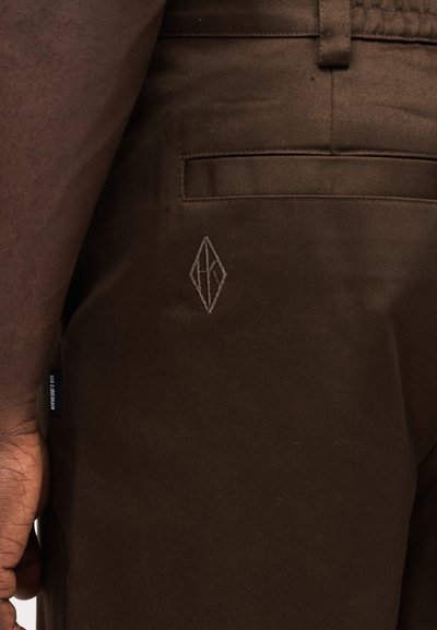 Brown cotton pants with a slim fit, featuring a back pocket and a small diamond-shaped logo embroidered near the pocket.