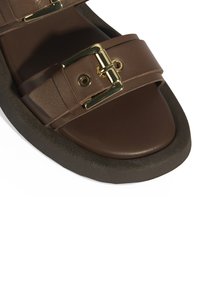 Sandale en cuir marron avec deux larges sangles et des accessoires en métal doré. Présente un bout arrondi et une épaisse semelle noire.