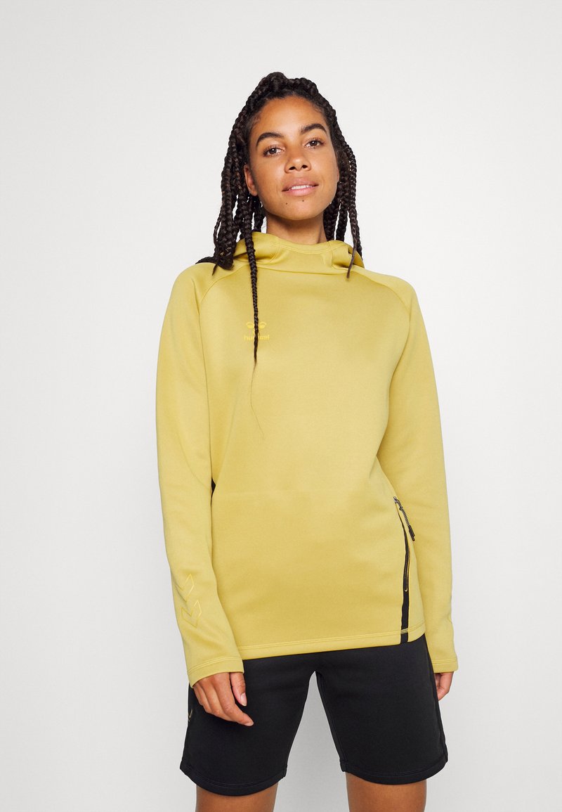 Jeune femme avec des cheveux tressés portant un sweat à capuche jaune et un short noir, se tenant devant un fond clair uniforme.