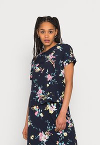 Camisa floral azul-marinho com mangas curtas; apresenta flores em tons de rosa, amarelo e verde-água; tecido macio; corte descontraído; combinada com uma saia a condizer.