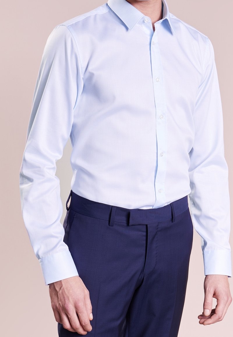 Homme portant une chemise bleu clair à manches longues boutonnée et rentrée dans un pantalon de costume bleu foncé, sur un fond uni.