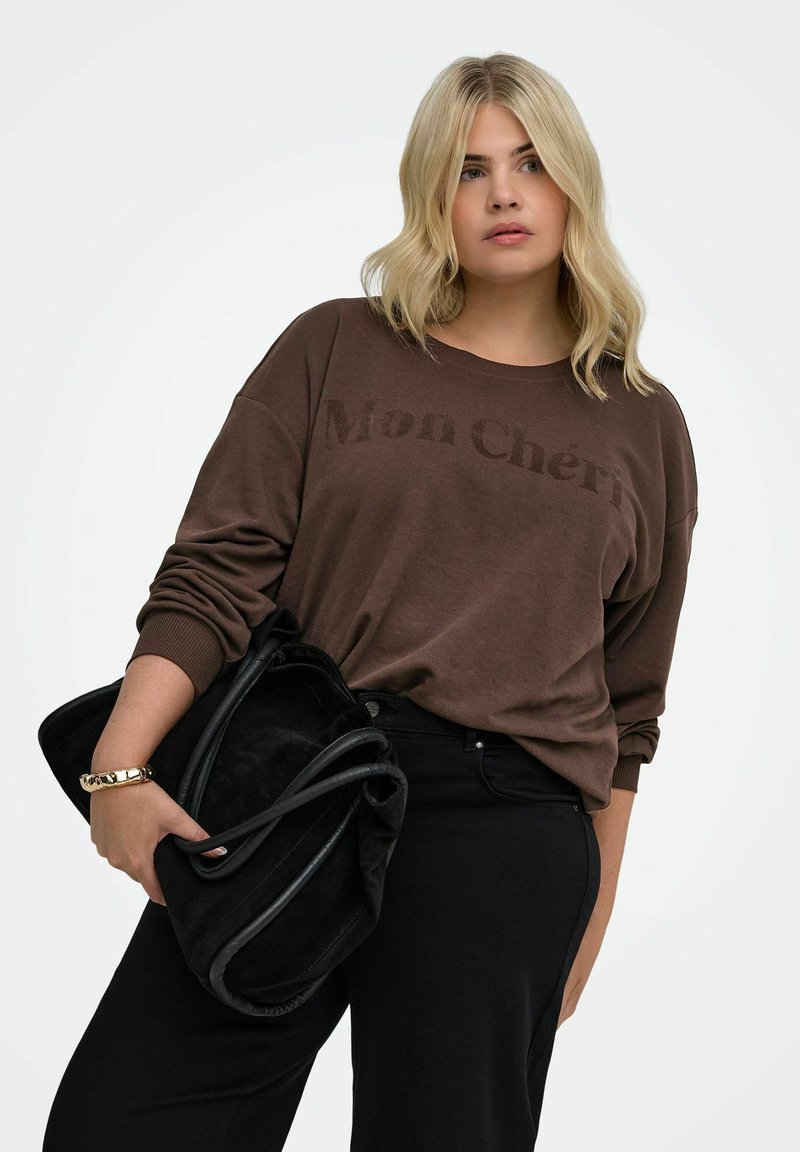 Femme blonde portant un sweat-shirt marron "Mon Chéri" et un pantalon noir, tenant un grand sac à main noir doux avec un bracelet doré au poignet gauche.