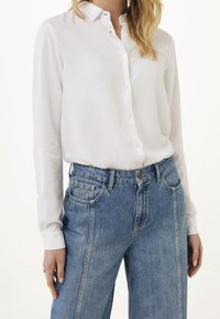 Kvinna som bär en vit skjorta med långa ärmar inskjuten i högmidjade blå denimjeans med synliga bältesöglor.