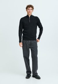 Schwarze Zip-Up-Trainingsjacke mit strukturiertem Finish, kombiniert mit lässigen, locker sitzenden dunkelgrauen Hosen und schwarzen Schuhen. Das Model steht vor einem hellen Hintergrund.