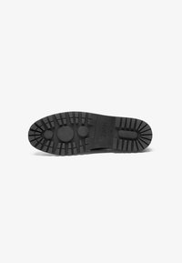 Suela de zapato de goma negra con superficie texturizada, que presenta surcos profundos y agarres circulares para mayor tracción. Logo visible cerca del talón.
