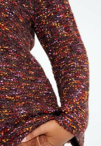 Stickad tröja med ett texturerat mönster som har flerfärgade fläckar i rött, orange, gult och lila på en mörkbrun bakgrund.