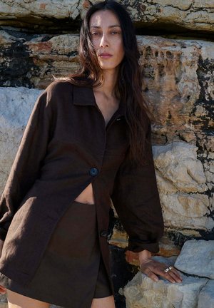 Mujer con el pelo largo y oscuro, vistiendo una chaqueta marrón oscuro con botones y falda, apoyada contra una pared rocosa con textura al aire libre.
