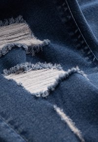 Primer plano de una tela de mezclilla azul con tres parches rasgados horizontales que muestran hilos blancos debajo.