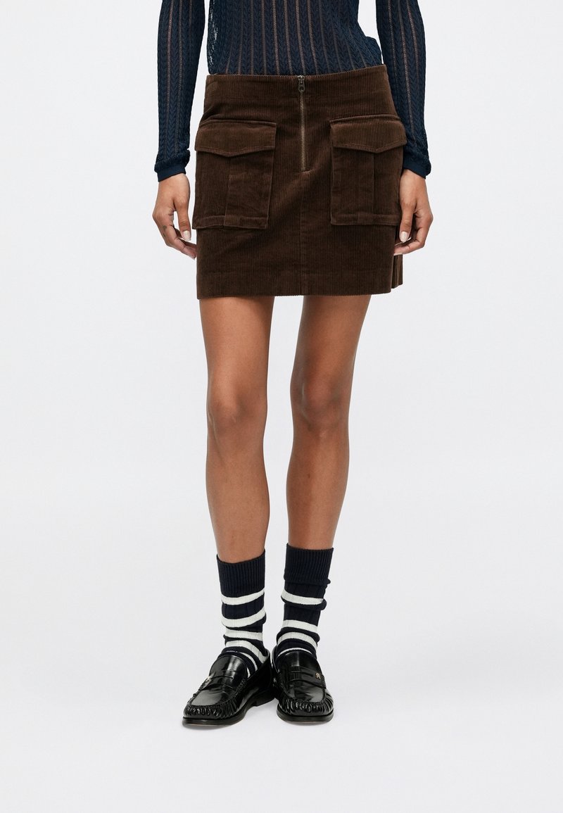 Femme portant une mini-jupe en velours côtelé marron avec des poches à l'avant, des mocassins noirs et des chaussettes rayées noir et blanc sur fond blanc.