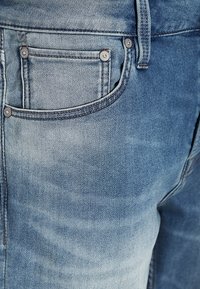 Los vaqueros de denim azul claro presentan un bolsillo trasero estándar con un detalle de remache, una textura desgastada y un sutil pespunte en las costuras.
