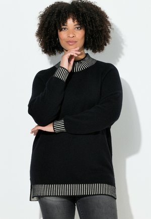 Kvinde med krøllet hår, iført en sort sweater med hvidt stribet krave, manchetter og kant, poserer med den ene hånd nær sin hage.