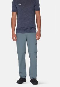 T-shirt pour homme bleu marine avec logo, associé à un pantalon gris clair avec zip amovible. Le pantalon a une texture lisse et présente un emblème noir sur la cuisse.
