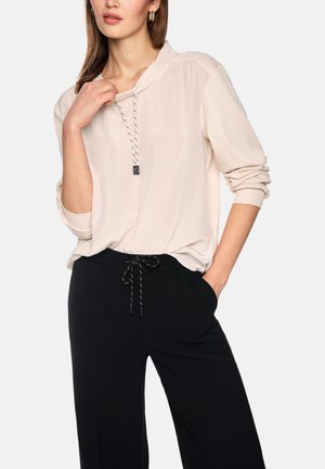 Woman wearing a beige long-sleeve blouse with drawstring neckline and black pants with drawstring waist, posing with one hand in pocket.