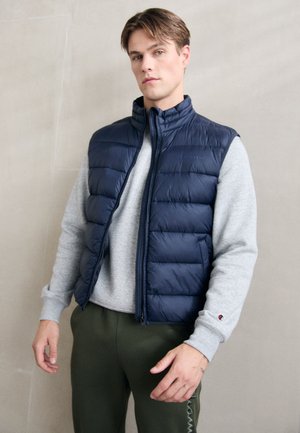 Navy puffer vest with a high collar, quilted design, and front zipper, worn over a light gray sweatshirt and olive green pants.