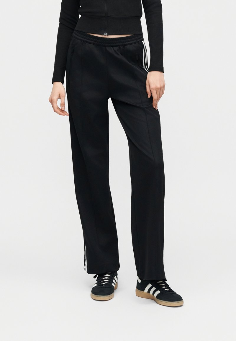 Femme portant un pantalon de sport noir avec des bandes blanches sur les côtés et des baskets noires à rayures blanches, debout devant un fond uni.