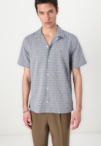 Camicia a maniche corte con bottoni in tessuto azzurro chiaro con un motivo sottile. Presenta un colletto alla canadese e bottoni bianchi, abbinata a pantaloni beige.