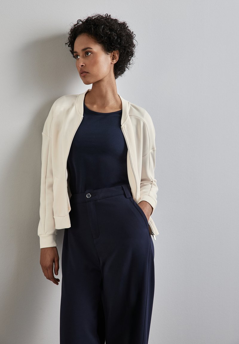 Una chaqueta bomber de crema clara sobre una camiseta navy, combinada con pantalones de tiro alto en color navy. Tejido suave con un corte relajado y detalles de diseño mínimos.