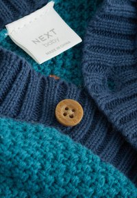 pleten pulover s turkiznim delom in temno modrim rebrastim ovratnikom, z lesenim gumbom z napisom "NEXT" in pripetim navodilom za nego.