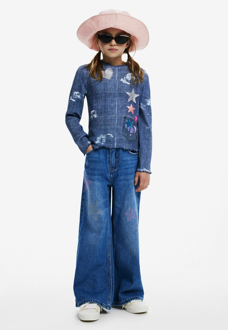 Džínová bunda so zničenou textúrou, farebné hviezdne aplikácie, skombinovaná s širokými modrými džínsami s trblietavými hviezdami. Biele tenisky a ružový vedierkový klobúk.