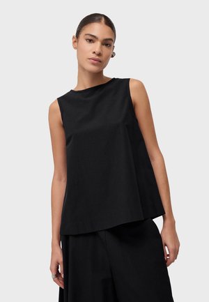 Žena s kosom zategnutom unatrag, odjevena u lepršavu crnu majicu bez rukava i crne hlače, stoji ispred jednostavne svijetle pozadine.