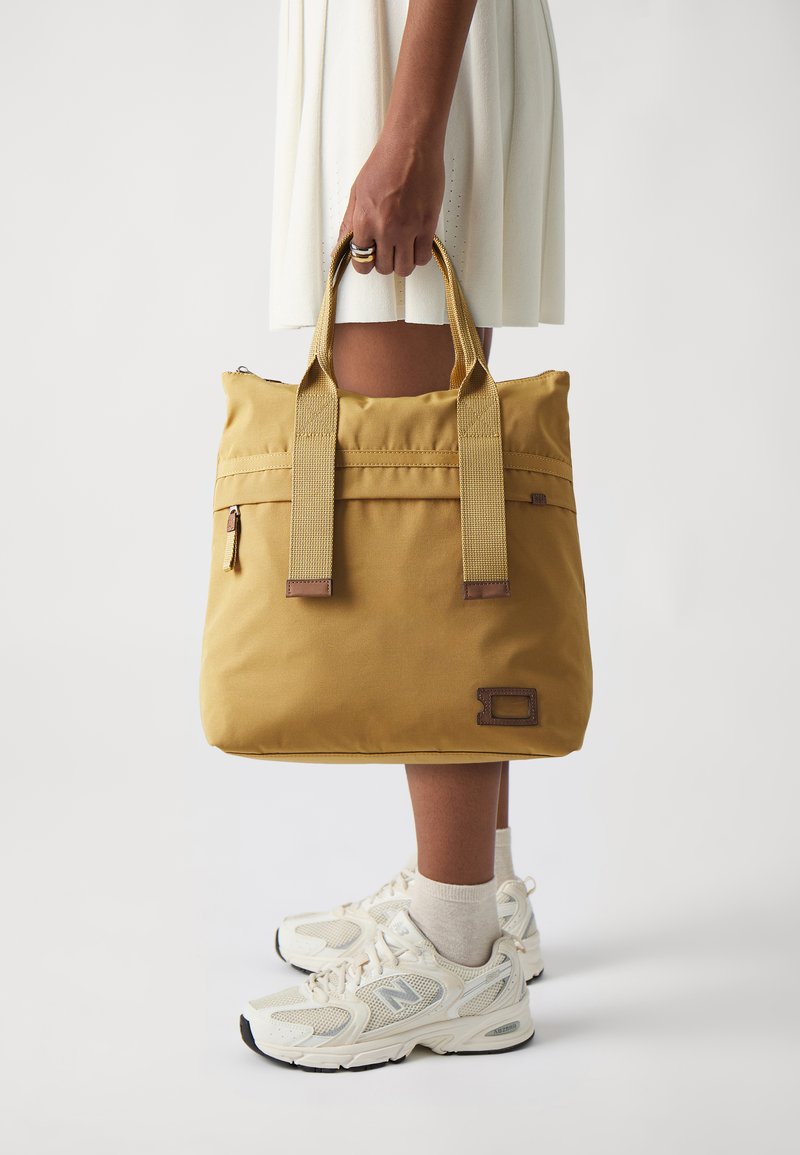 Yellow fabric tote bag with two wide handles, a zippered top, and a small front pocket. Features brown accents and logo detail.