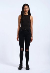 Musta hihaton toppi yhdistettynä mustiin repeytyneisiin kapeisiin farkkuihin, joissa kulutetut polvikoristeet, ja mustat urheilukengät. Yhtenäinen värimaailma.