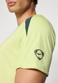 Camiseta deportiva de color verde lima con un acabado texturizado, que presenta acentos azul oscuro en el hombro y un logo negro de Nike en la manga.