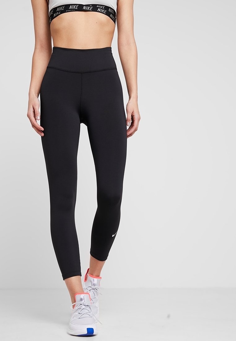 Femme portant un legging noir taille haute, un soutien-gorge de sport blanc Nike et des chaussures de sport blanches avec des touches rose et bleu, debout sur un fond uni.